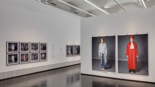 Ein Ausstellungsraum an dessen Wänden drei Bilder hängen, auf denen lebensgroß Menschen abgebildet sind. Auf dem vordergründigsten Bild das Foto einer Frau, die einerseits eine rote Richter-Robe trägt und andererseits eine Jeans und eine hellblaue Trainigsjacke.