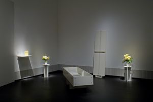 Dunkler Ausstellungsraum. In der Ecke des Raumes steht ein geöffneter weißer Sarg aus Holz. Links und rechts davon steht jeweils eine Blumenvase auf einem Sockel.