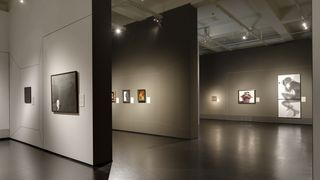 Einen Ausstellungsraum in dessen Mitte ein abgetrennter Bereich, in welchem Gemälde hängen. An den anderen Wänden ebenfalls Gemälde und Fotografien. 