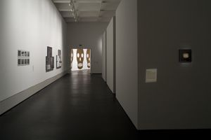 Blick in einen Ausstellungsraum. Rechts hängen Fotografien und Bilder an der weißen Wand. Im Hintergrund eine beleuchtete Wand auf der drei ca. zwei Meter hohe Totenköpfe abgebildet sind.
