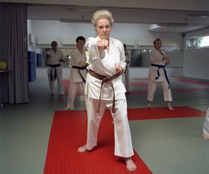 Eine alte Frau in Karatekleidung in einer Karatestellung steht auf einem roten Teppich. Im Hintergrund sind weitere Personen in der gleichen Haltung ebenfalls in Karatekleidung.