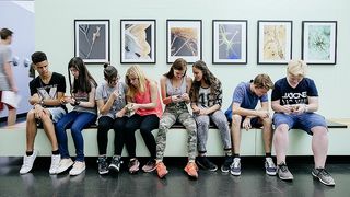 Eine Gruppe von weiblichen und männlichen Teenagern sitzt auf einer Bank vor einer Wand mit Bildern.