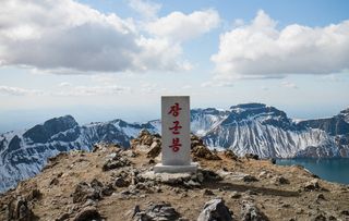 Auf einem Berggipfel steht ein helle Steintafel, auf der drei koreanische Schriftzeichen in rot eingelassen sind. Im Hintergrund sieht man einen kahlen, schneebedeckten Bergkessel und einen Bergsee. Der Himmel ist locker bewölkt.